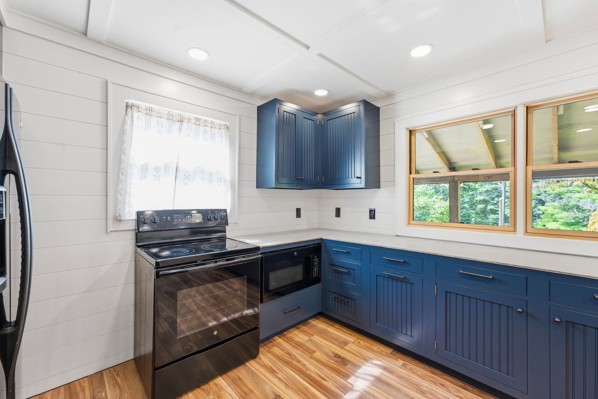 Kitchen with navy cabinets, black range, warm wood flooring, and windows overlooking trees.