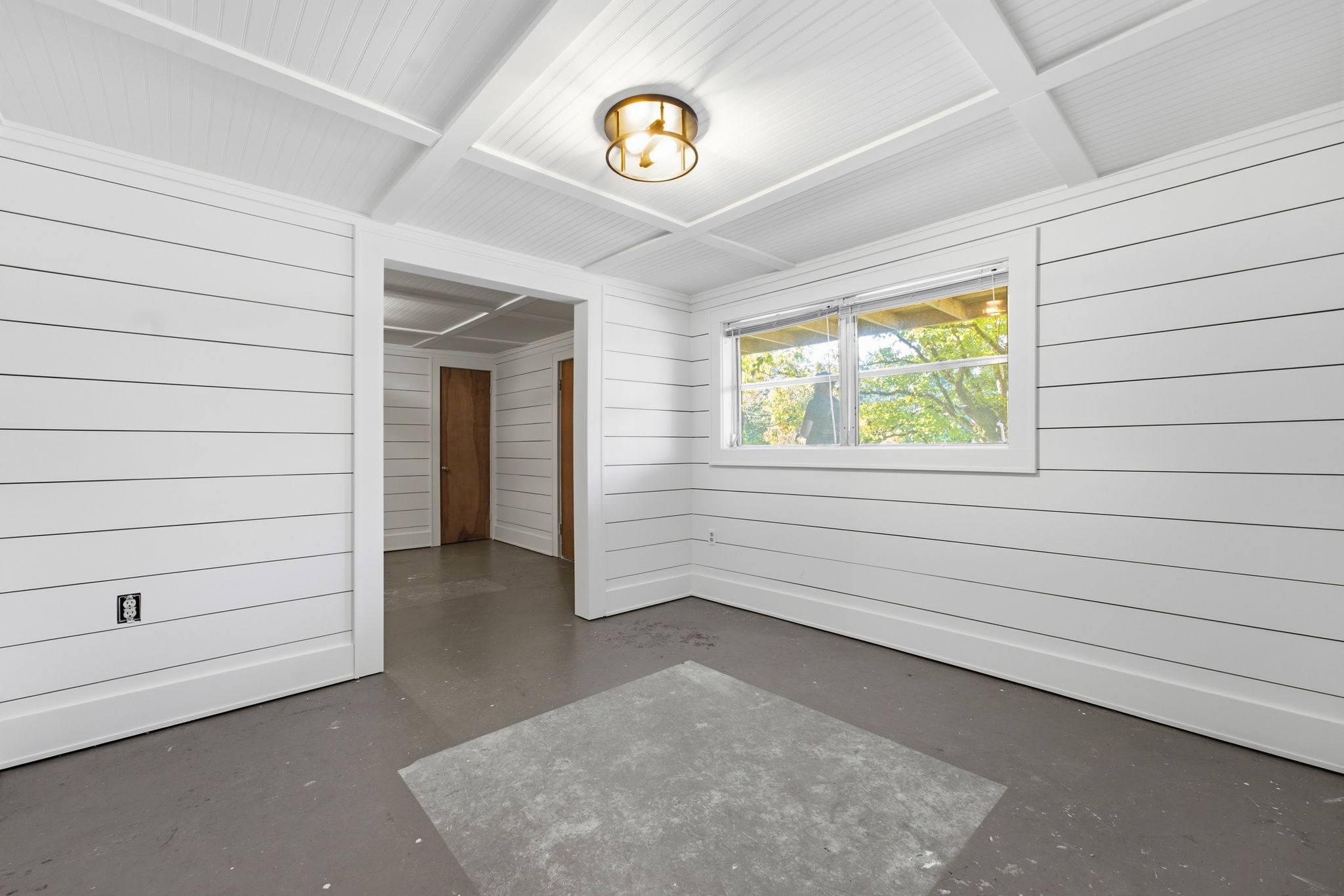 Empty room with white paneled walls, a vaulted beam ceiling, and a window overlooking trees.