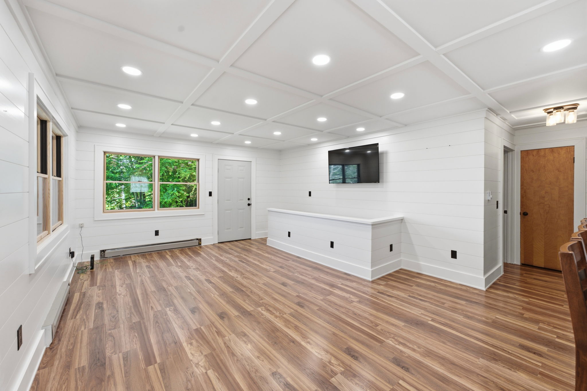 Bright white-paneled room with a coffered ceiling, wood-look floors, large windows, and a wall-mounted TV.