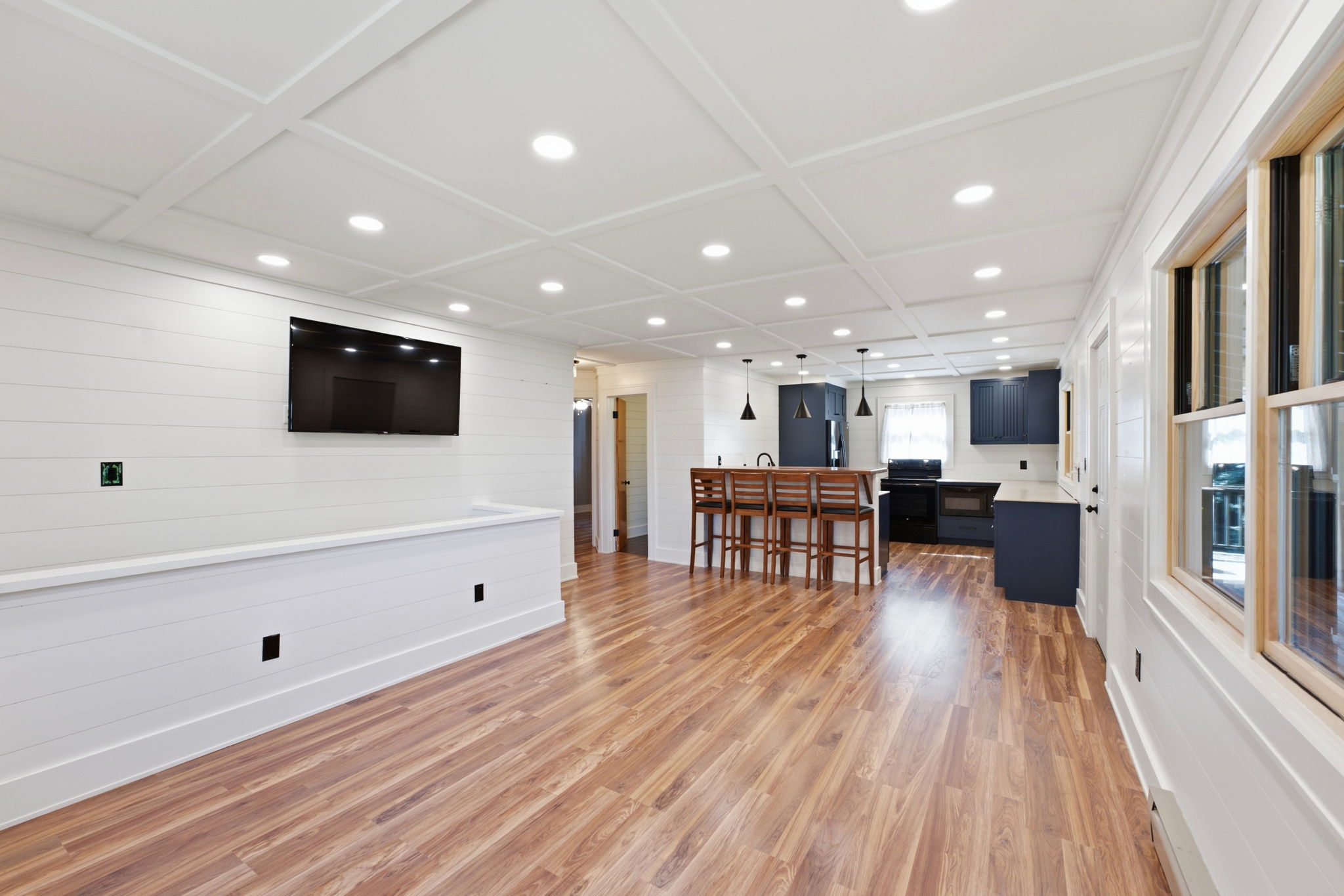 Open-plan kitchen with a wood-floored living area, coffered ceiling, island seating, and dark blue cabinets.