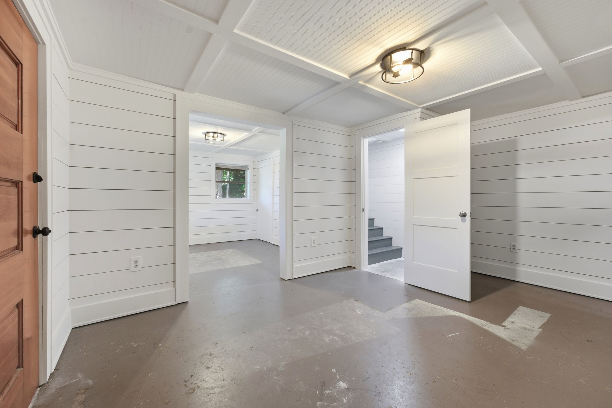 Empty interior with white plank walls, a vaulted beamed ceiling, concrete floor, and stairs through an open doorway.