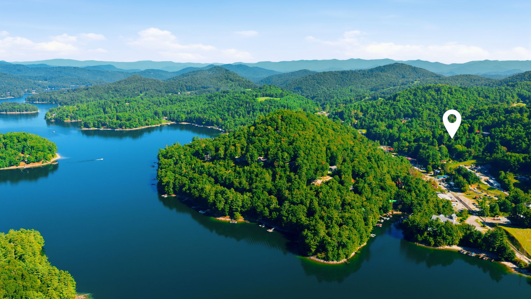 Aerial view of a wooded lakeside property marked by a pin, with surrounding mountain ridges.