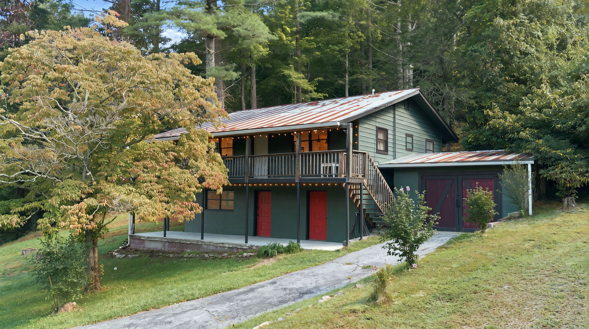 Green two-story home with a weathered metal roof, raised deck, and attached garage among trees.