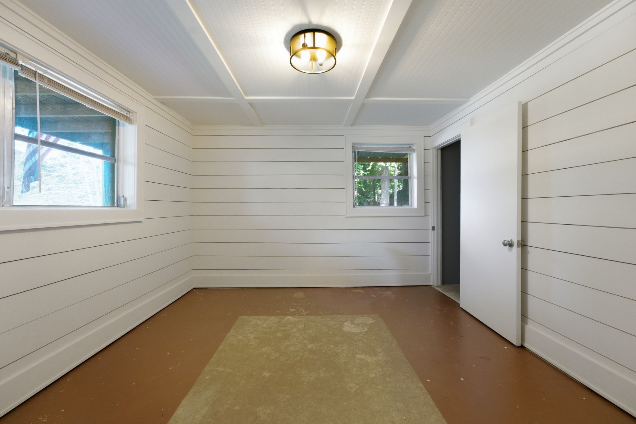 Empty room with white plank walls, a coffered ceiling, two windows, and a patchy brown floor.