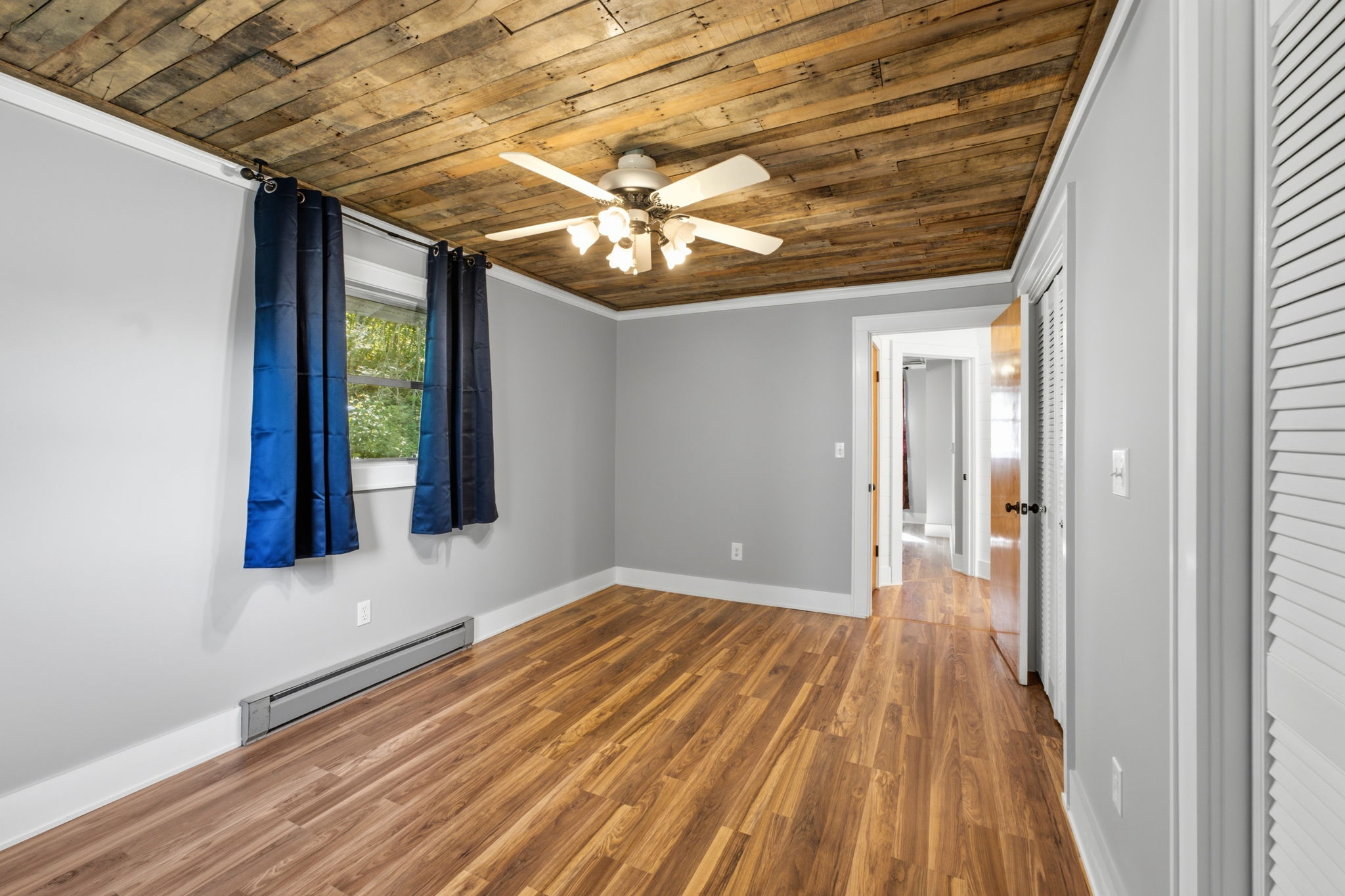 Empty bedroom with a wood-plank ceiling, wood-look floors, and blue curtains beside the window.