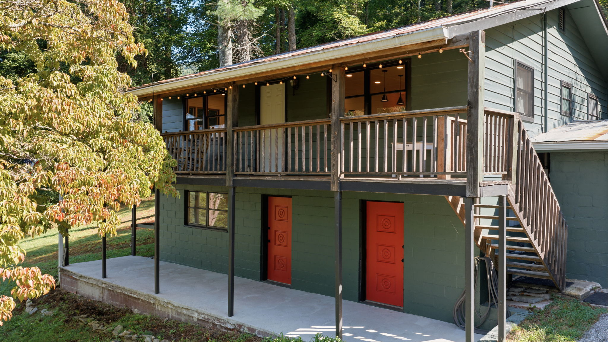 Green house with a wooden upper deck, exterior stairs, string lights, and bright red doors.