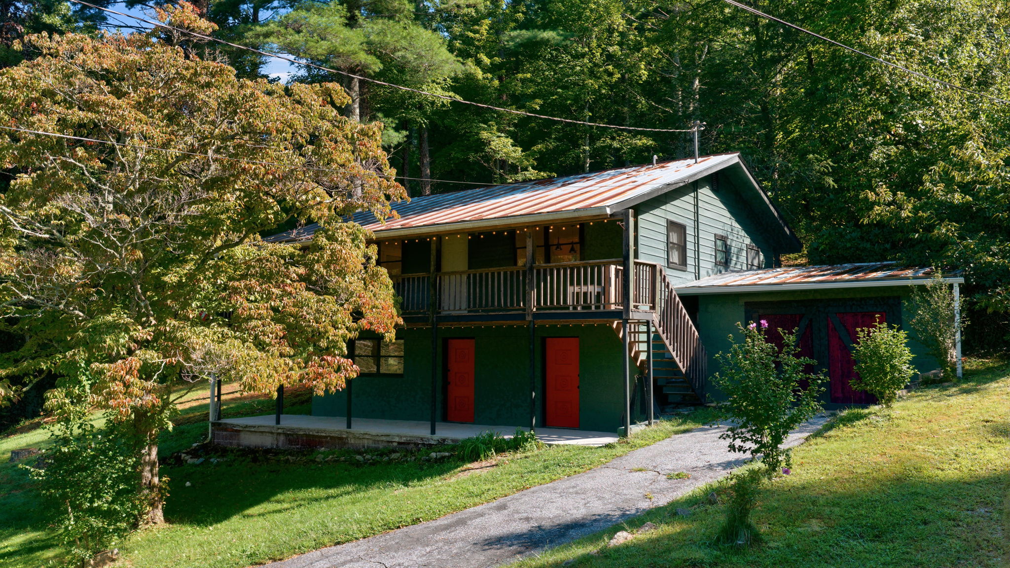 Wooded two-story green home with a raised deck, weathered metal roof, and red doors.