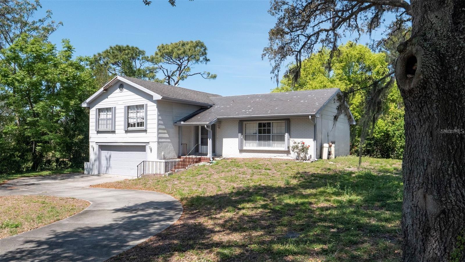 Light-colored two-story house with an attached garage and curved driveway beneath mature trees.