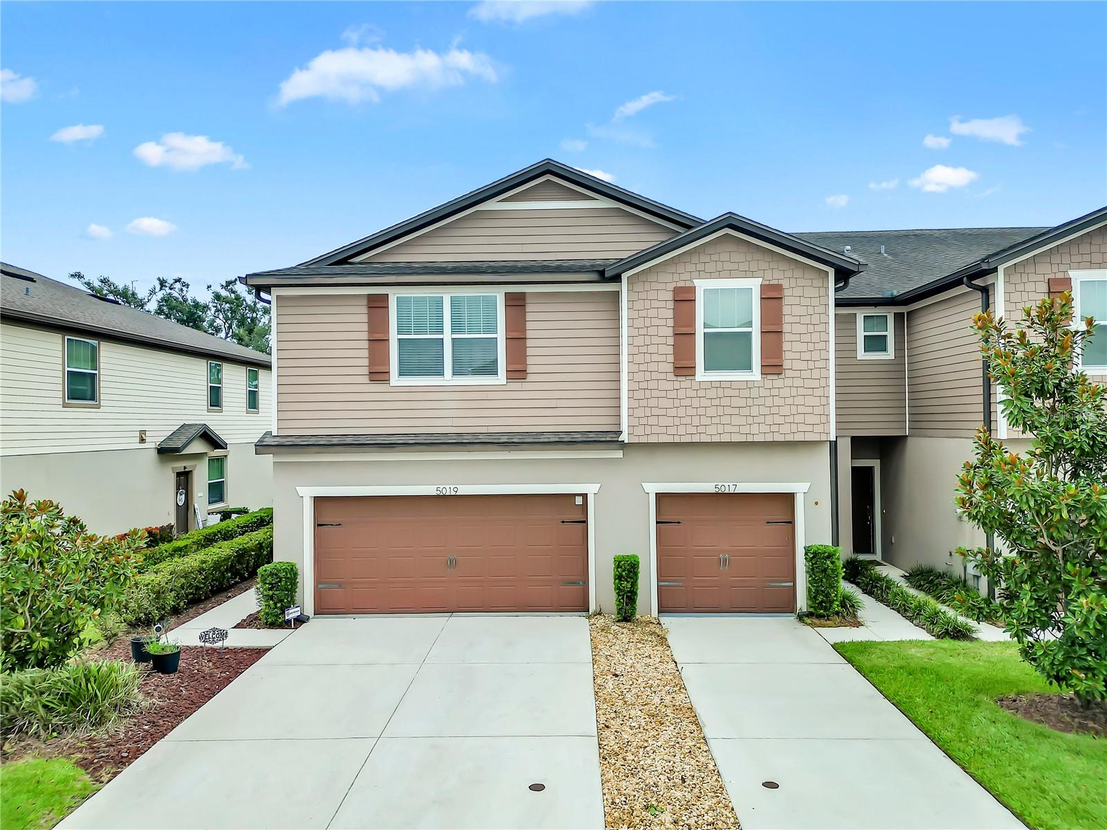 Two-story tan townhouse with shingle accents, paired brown garage doors, and landscaped front yards.