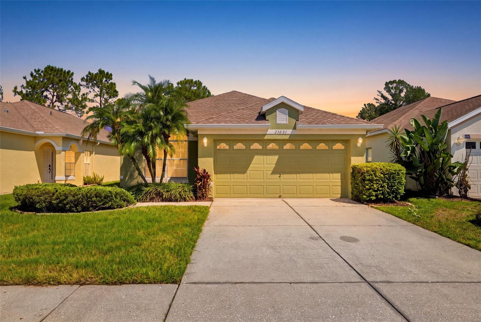 Single-story tan stucco home with a two-car garage, palm trees, and a wide driveway at sunset.