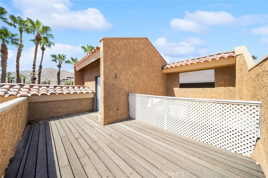 Wood-plank deck with white lattice railing, tan stucco walls, and palm trees in the distance.