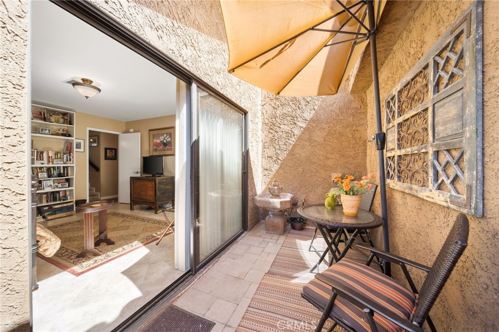 Private patio with an oversized tan umbrella, bistro table, potted flowers, and sliding doors to the living room.