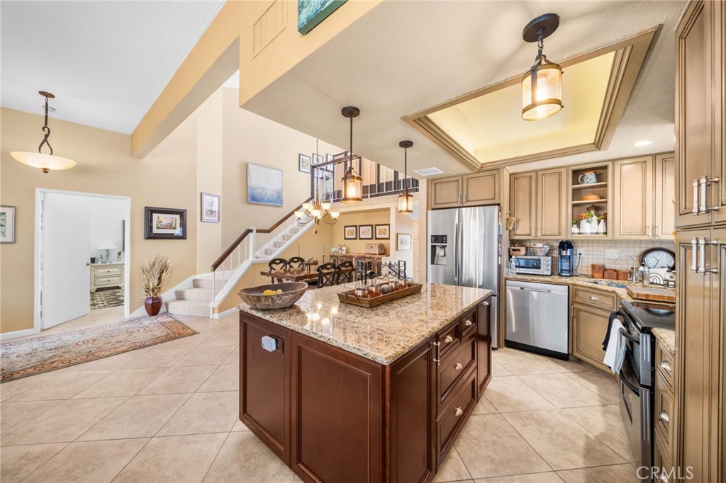 Open-plan kitchen with a granite island, tan cabinetry, stainless steel appliances, and a staircase.