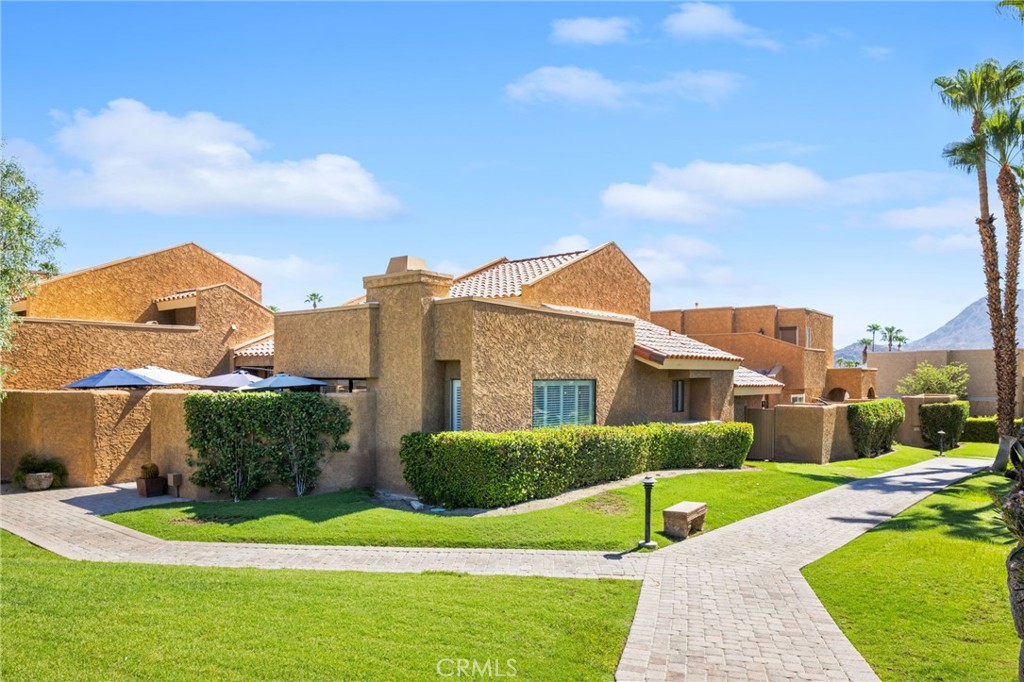Tan stucco home with tiled roofs, clipped hedges, a paved walkway, and palm trees.