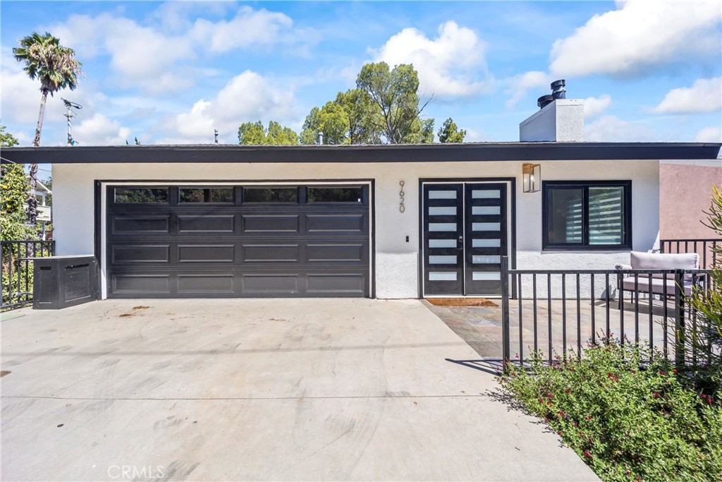 Modern single-story home with black garage door, wide concrete driveway, and patio seating.