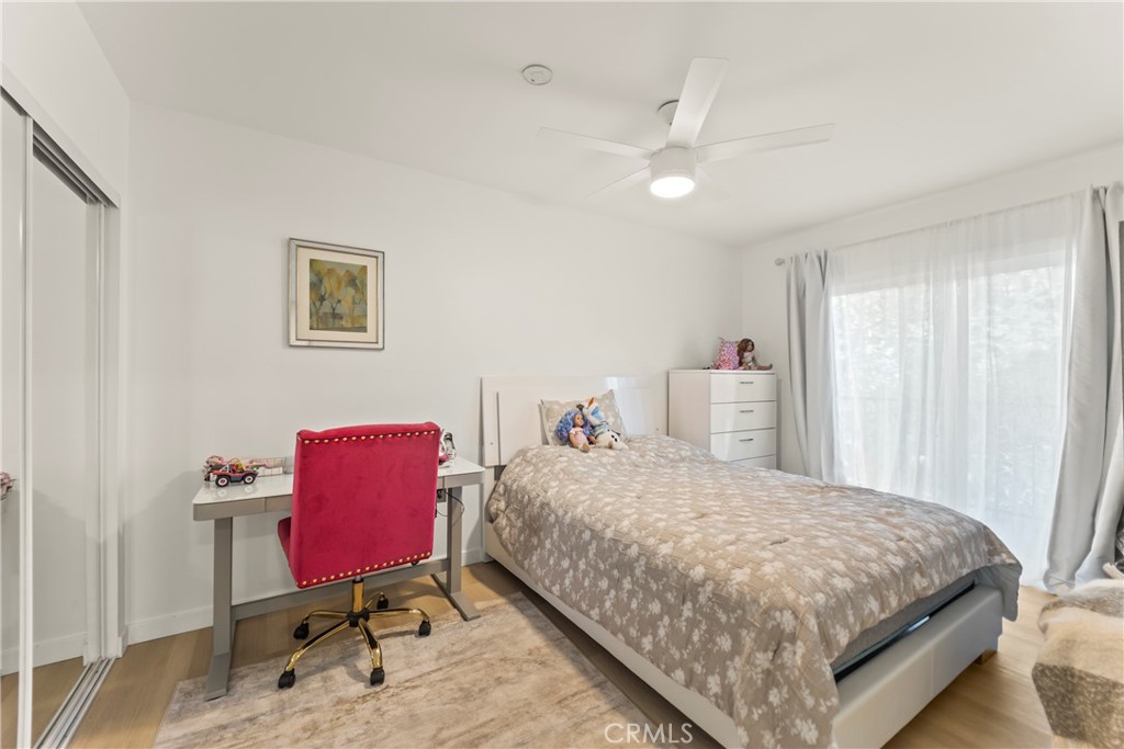 Furnished bedroom with a bed, desk, and bright red chair beside large curtained windows.