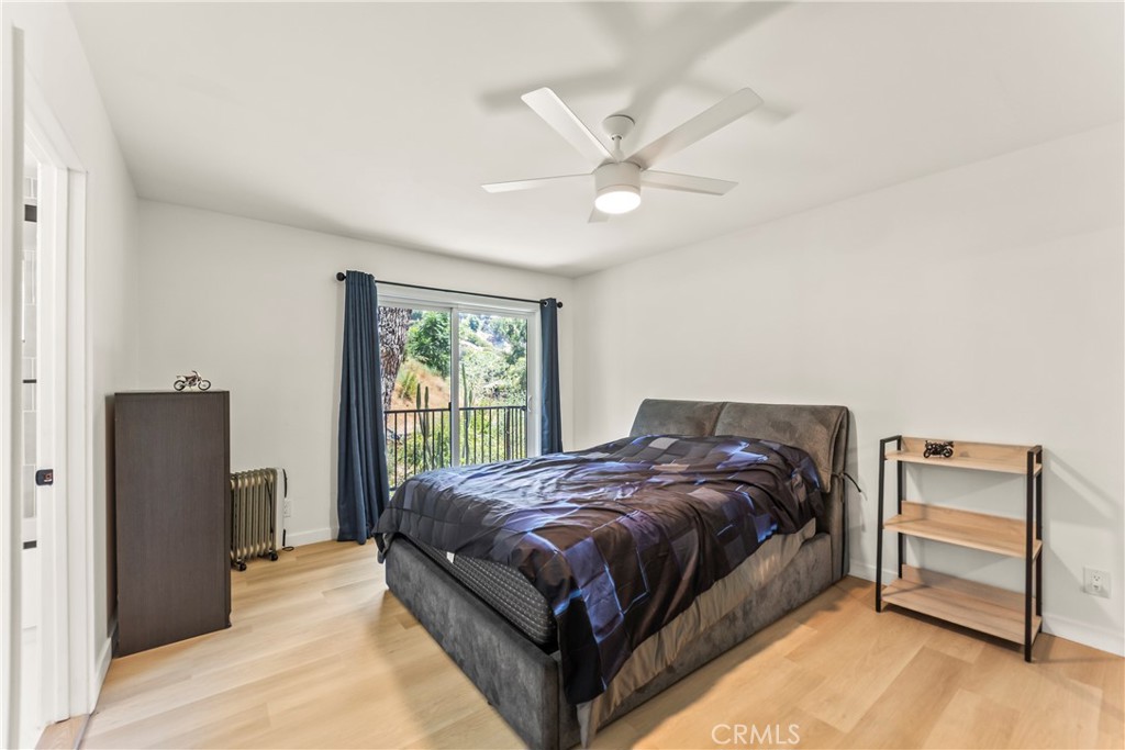 Bedroom with a gray upholstered bed, ceiling fan, light wood floors, and a balcony door overlooking greenery.