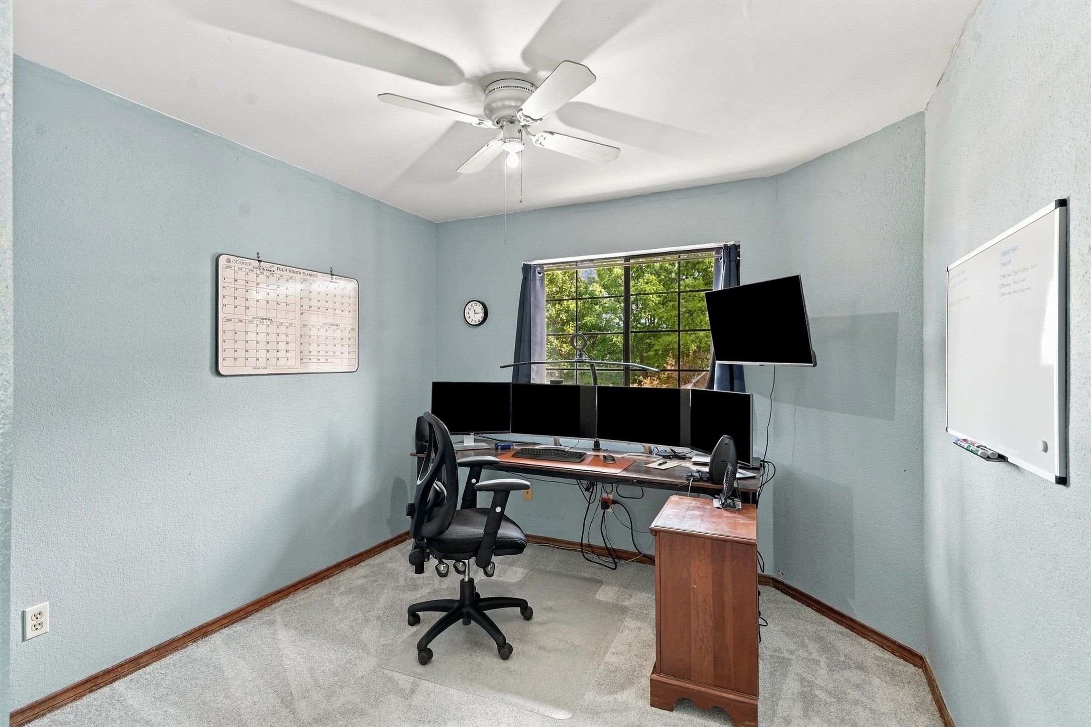 Home office with a multi-monitor workstation, wooden desk, and window overlooking trees.