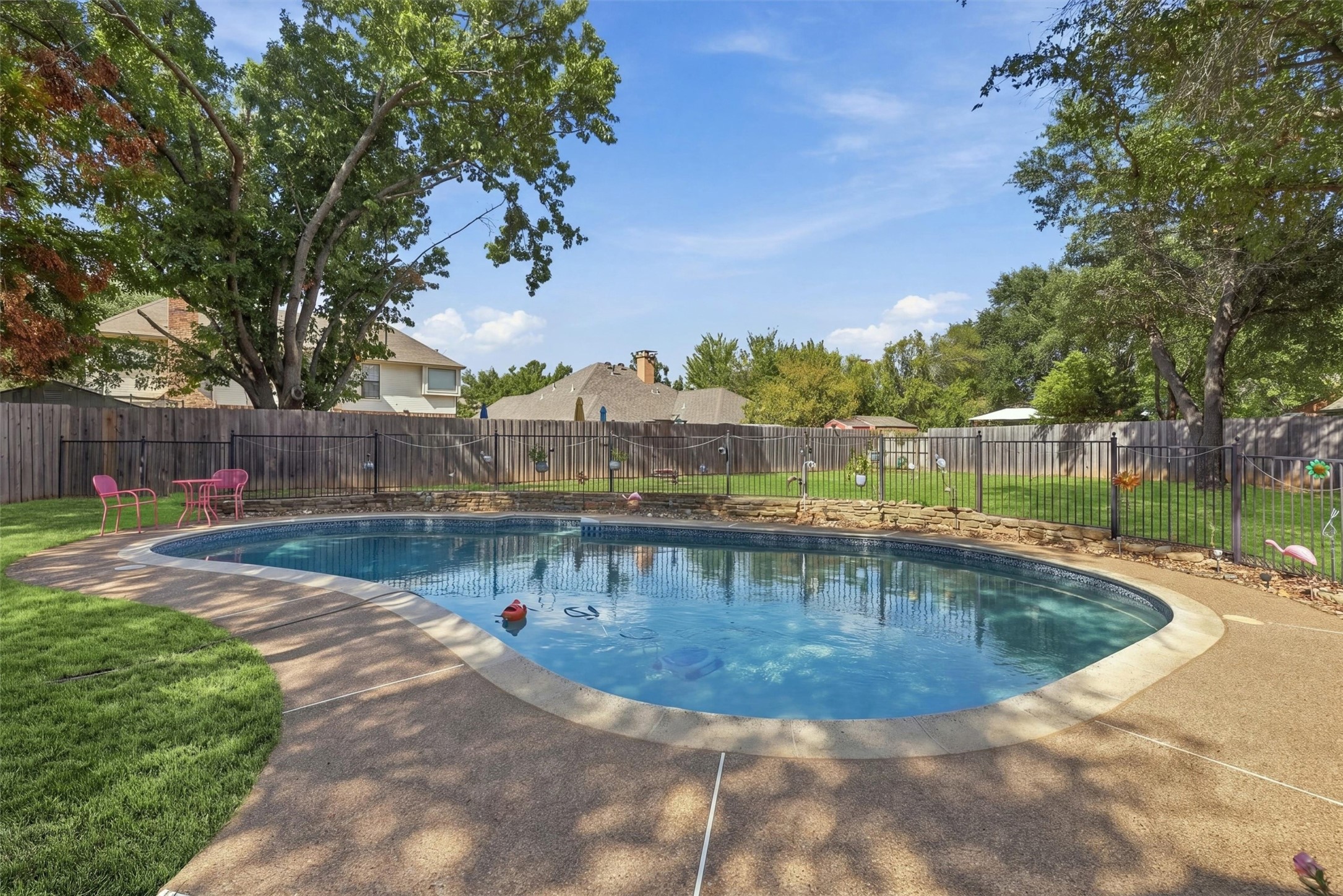 Backyard swimming pool with a curved deck, fenced lawn, and mature shade trees.