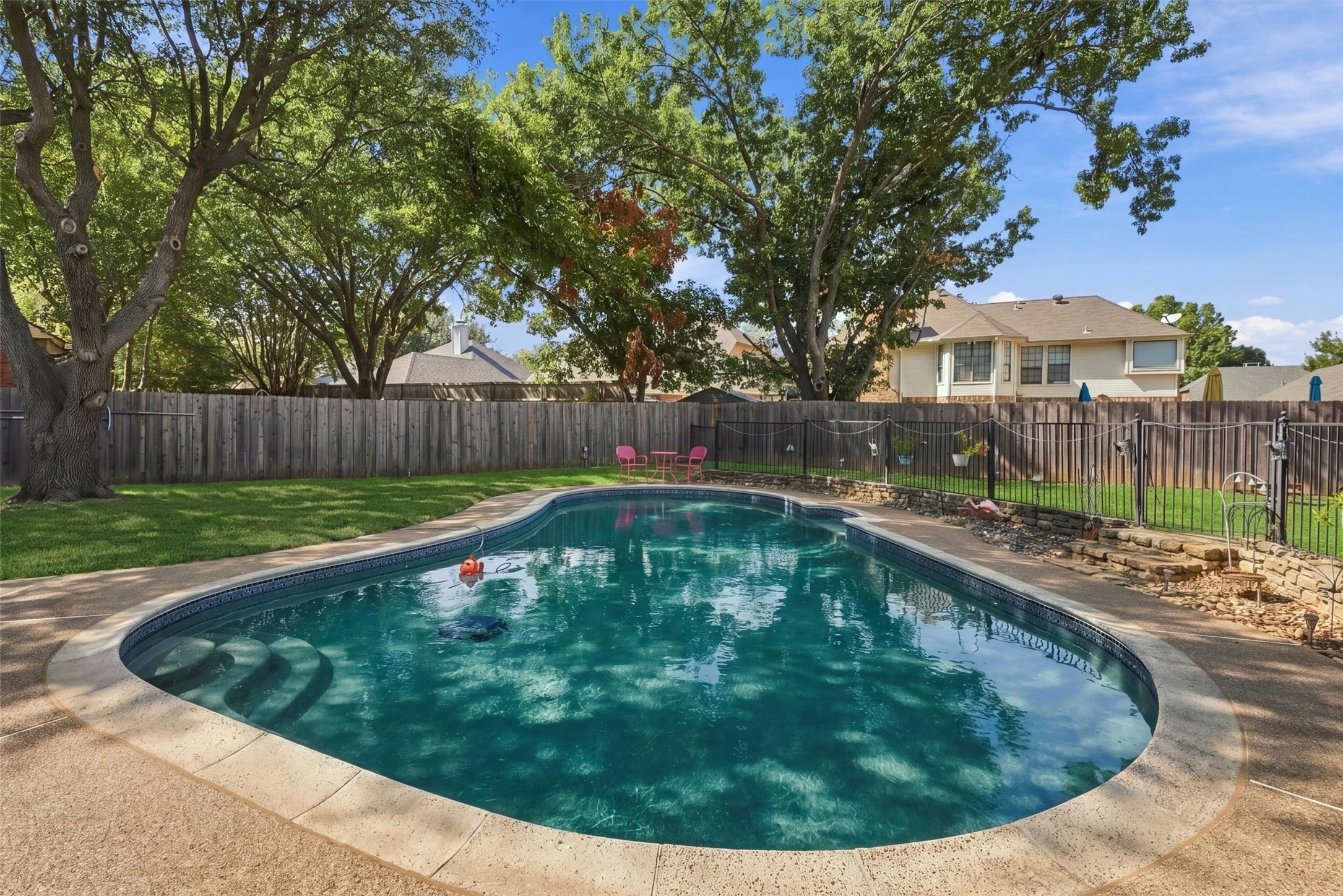 Backyard swimming pool with curved steps, mature shade trees, fenced lawn, and pink patio chairs.