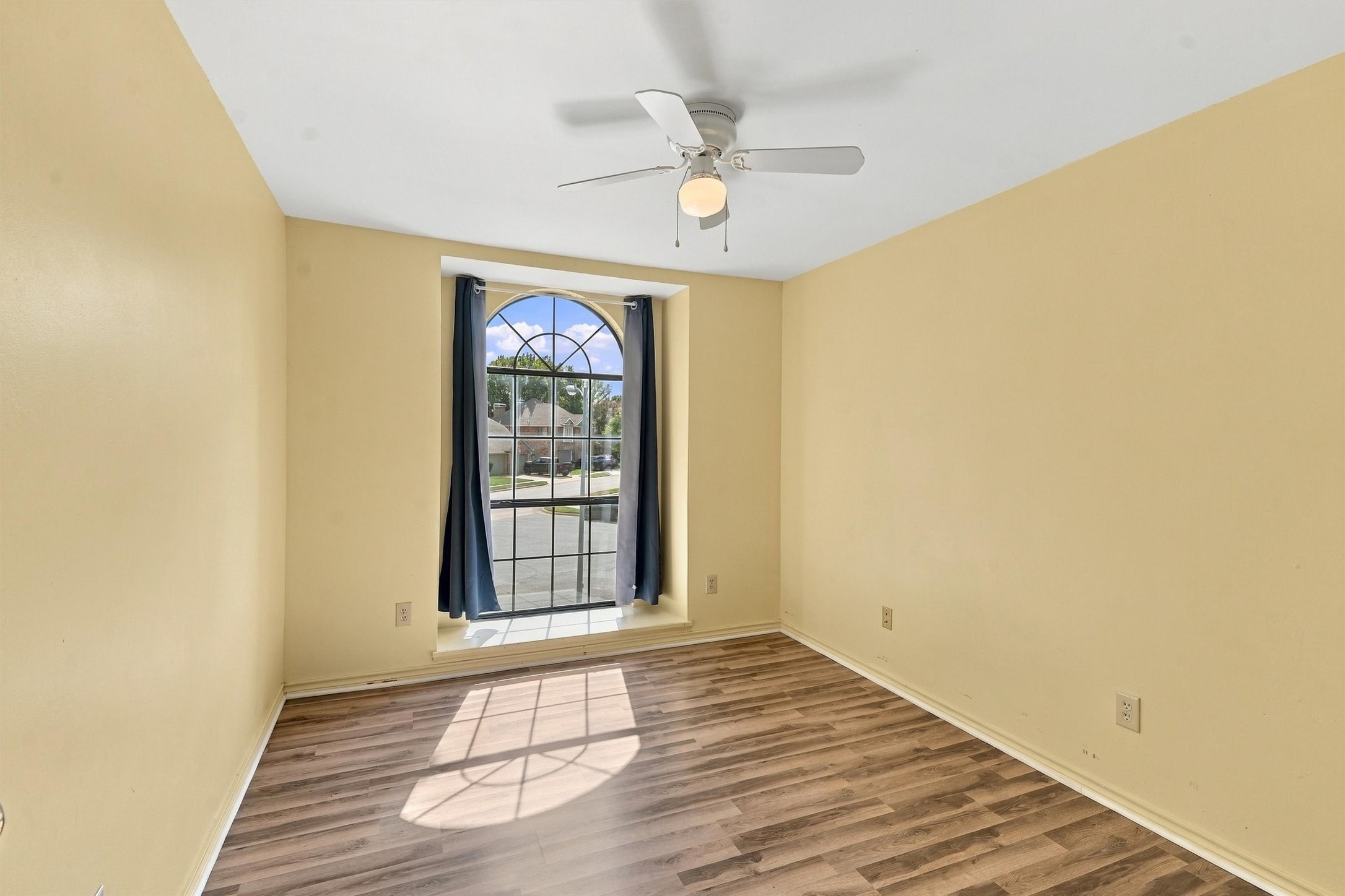 Empty room with wood-look flooring, a curtained arched window, and a ceiling fan.