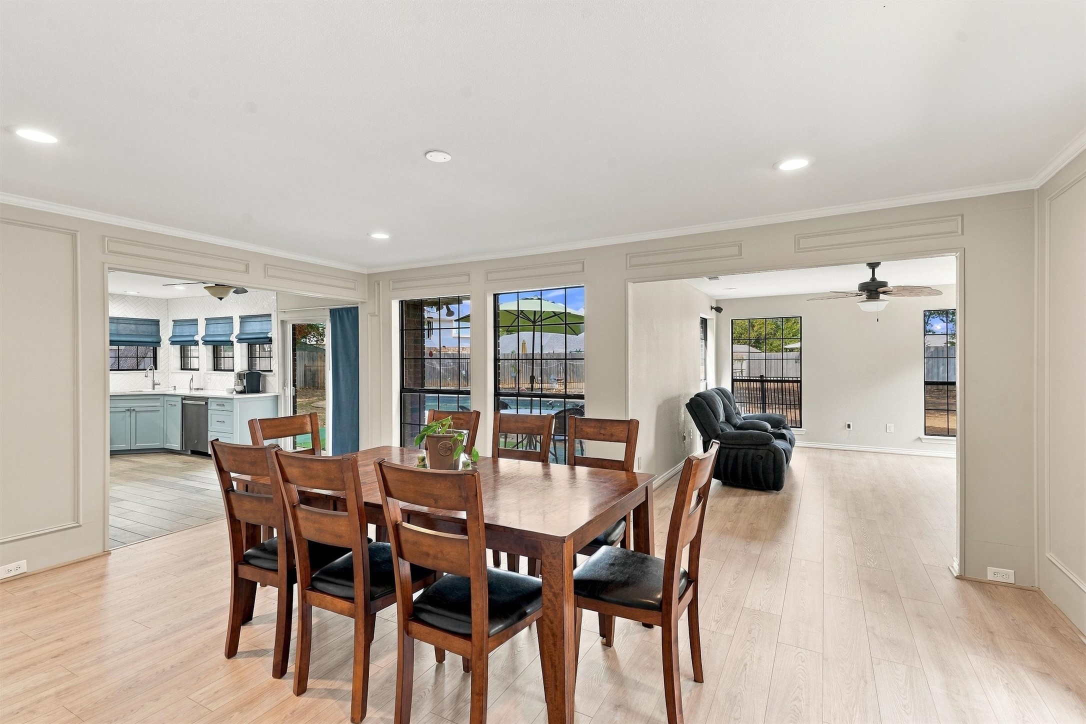 Dining room with a wooden table, adjoining blue kitchen and living area, and windows overlooking a pool.