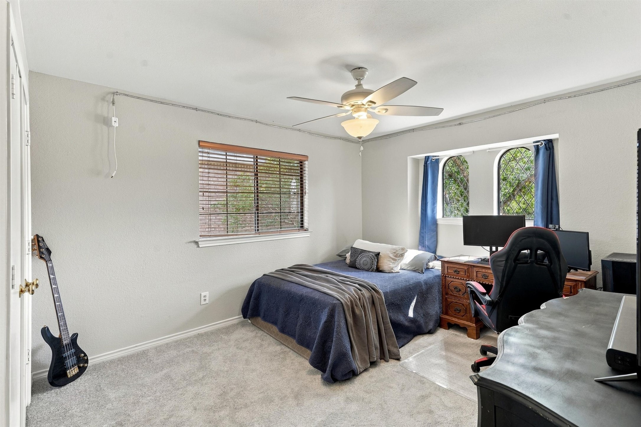 Bedroom with a blue-covered bed, dual-monitor gaming desk, and arched windows with blue curtains.