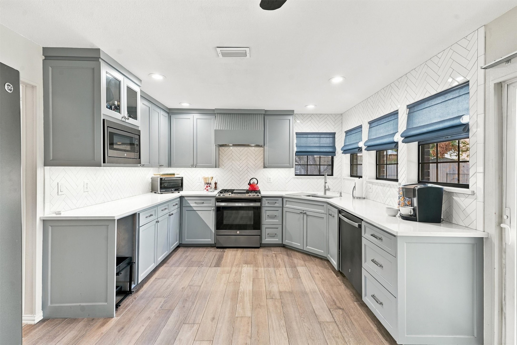 Gray-cabinet kitchen with white herringbone tile, blue window shades, and wood-look floors.