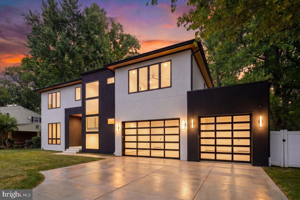 Modern two-story home with two illuminated garage doors and a broad driveway at dusk.
