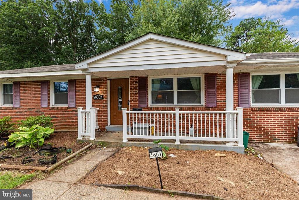 Red brick ranch home with a white-railed porch, burgundy shutters, and mature trees.