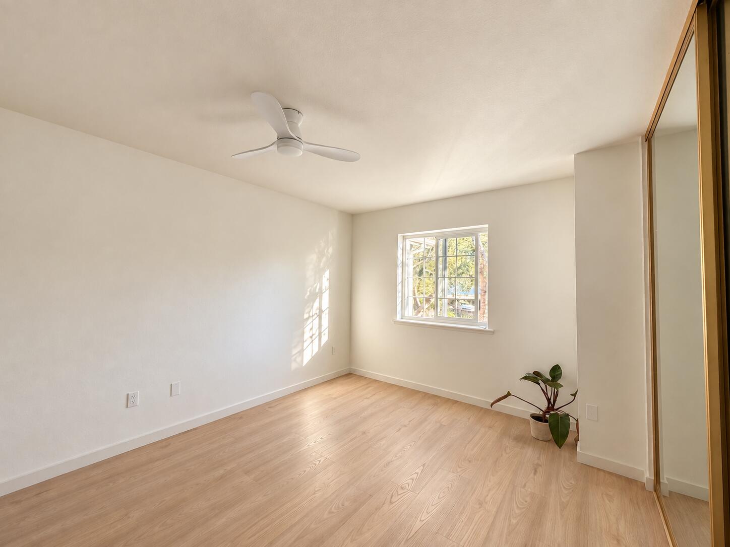 Empty bedroom with light wood floors, mirrored closet doors, and sunlight streaming through a window.