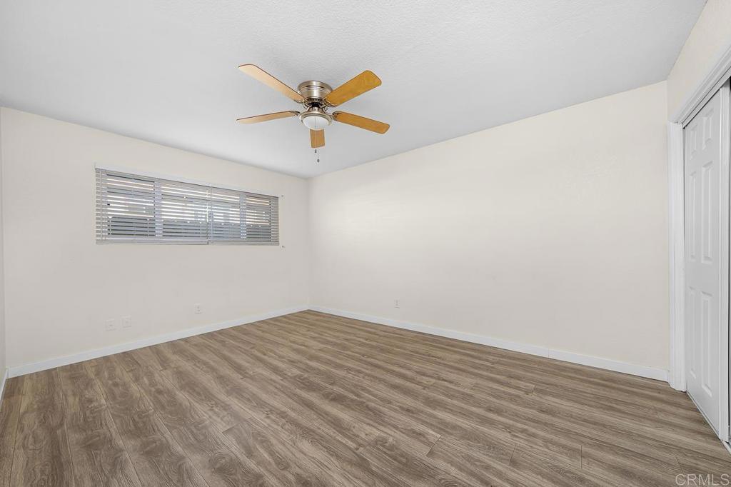 Vacant bedroom with wood-look flooring, ceiling fan, wide window with blinds, and closet doors.