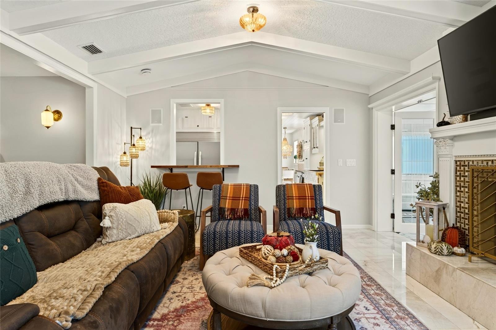 Staged living room with vaulted beamed ceiling, patterned armchairs, round ottoman, and fireplace with mounted television.