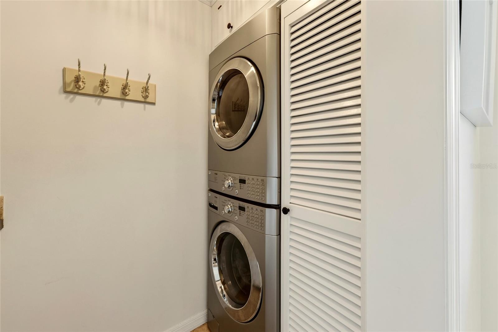 Stacked stainless steel washer and dryer beside a white louvered closet door.