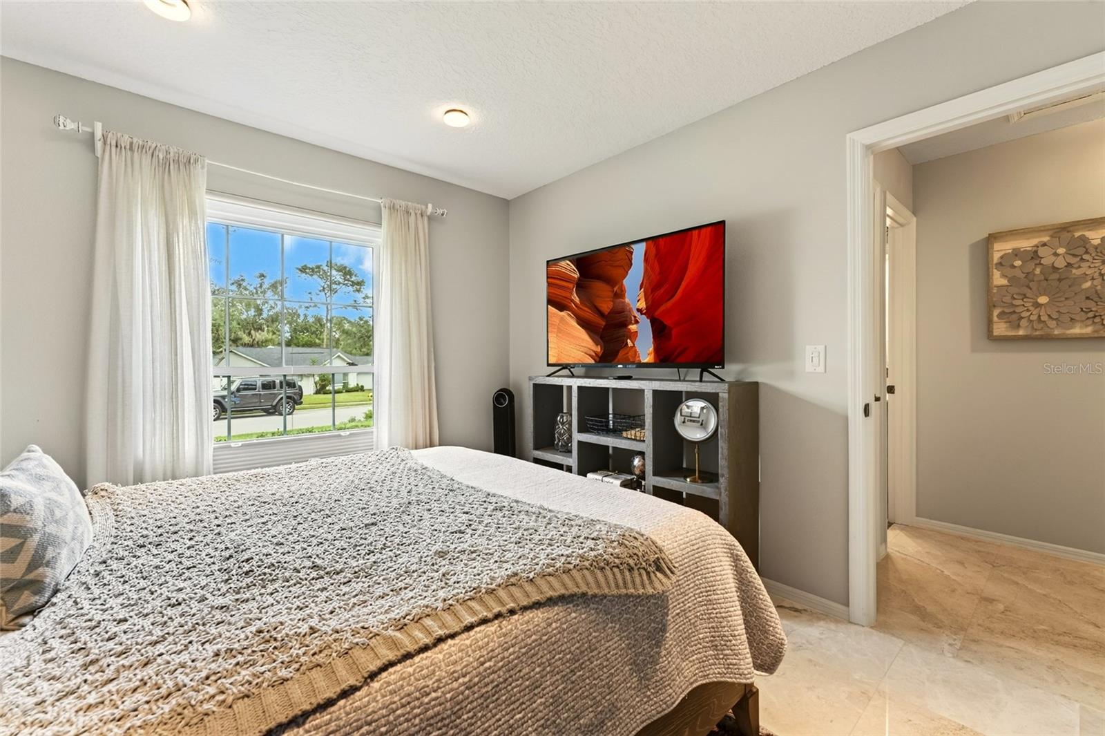 Furnished bedroom with a beige quilted bed, curtained street-facing window, and television on a console.