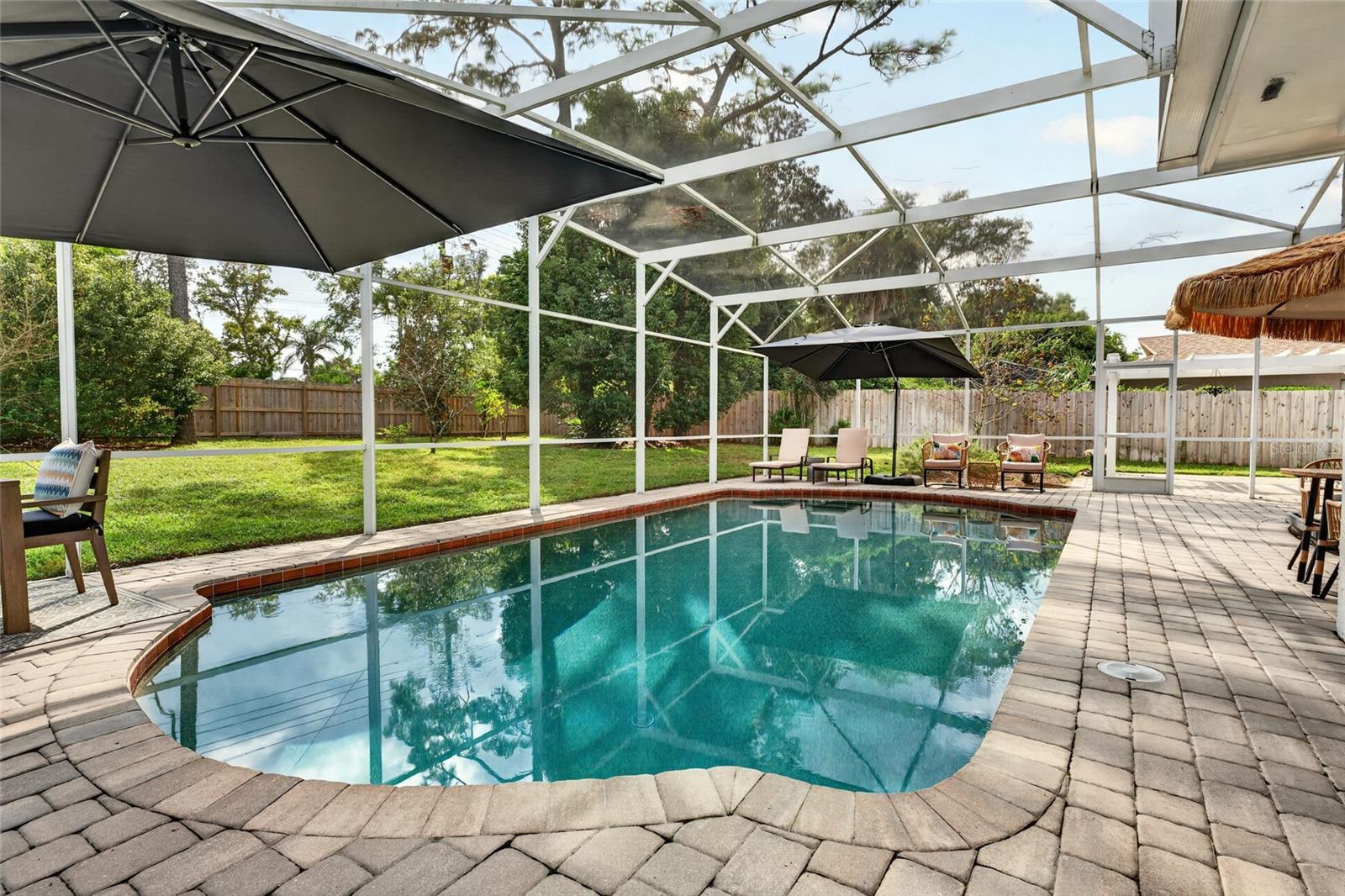 Screened-in backyard pool with stone pavers, patio chairs, and umbrellas.