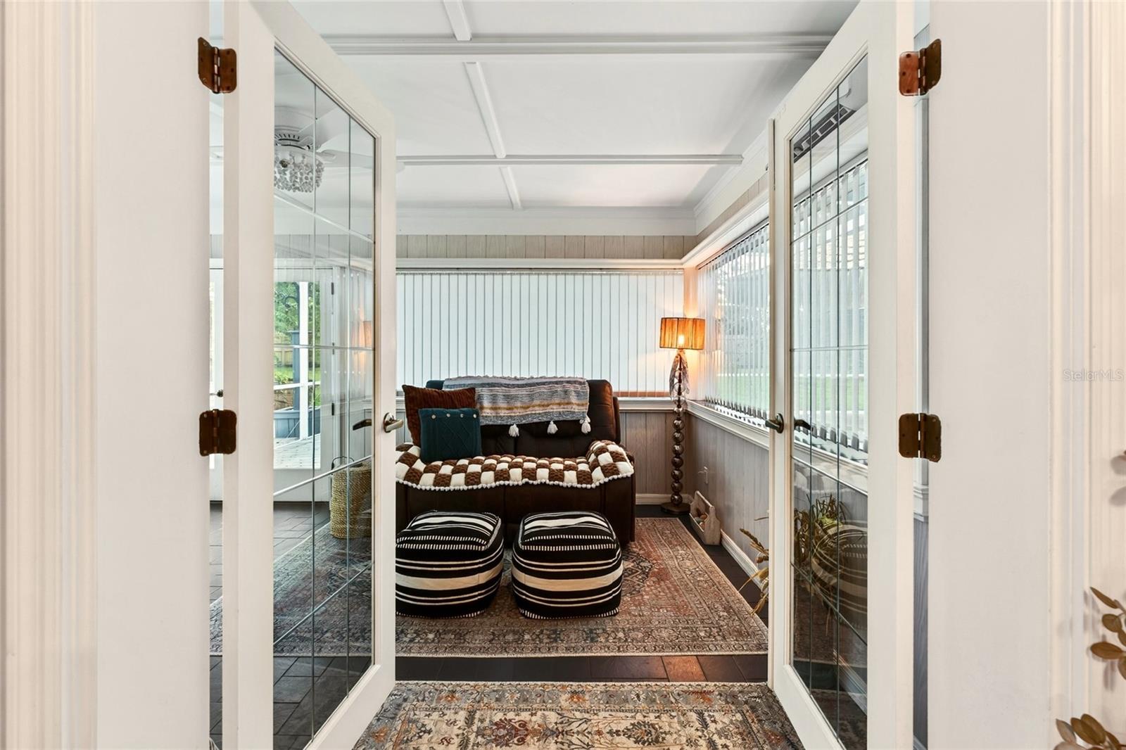 Furnished sunroom with a brown loveseat, striped ottomans, and wide windows with vertical blinds.