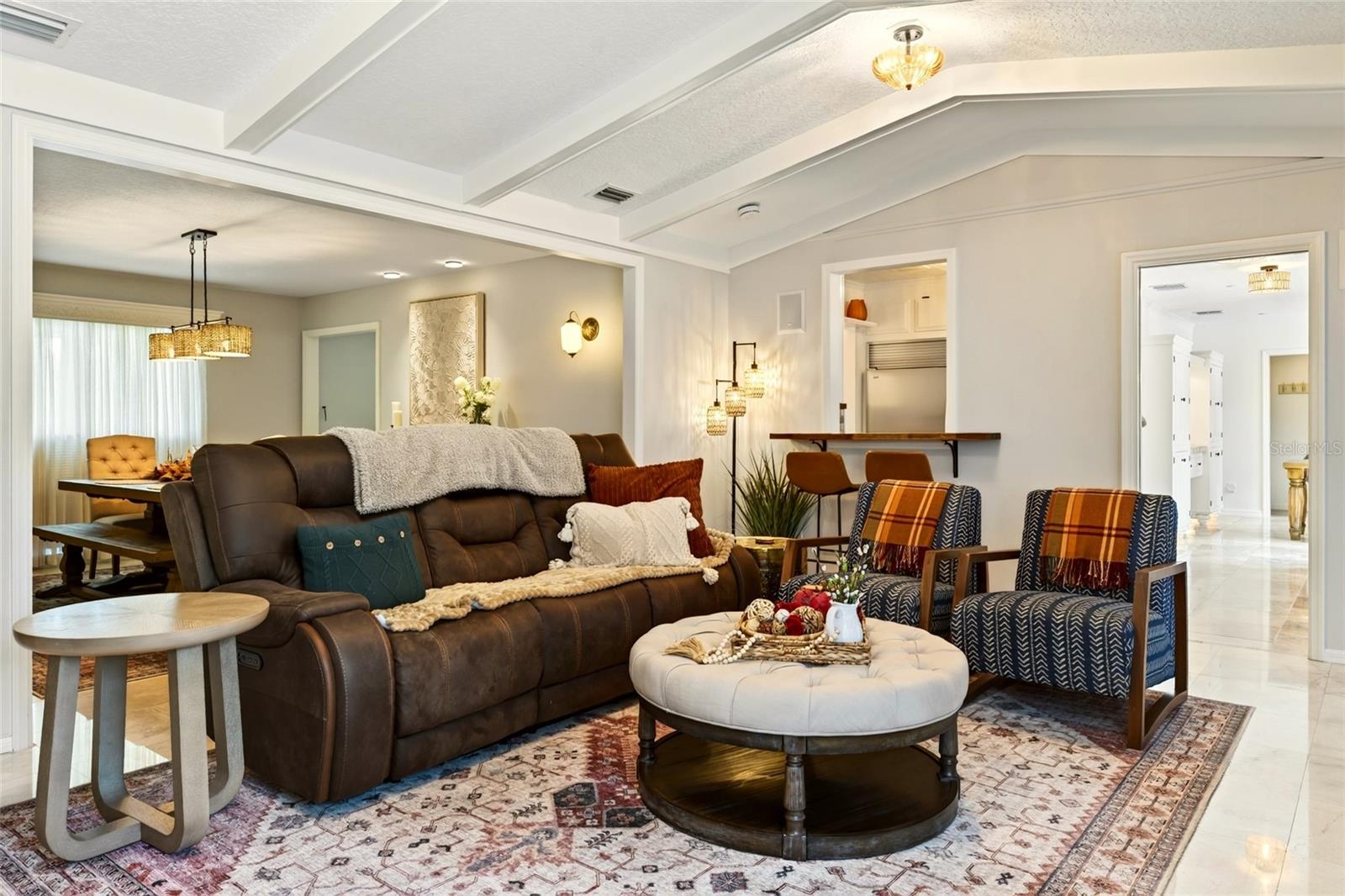 Living room with a leather sectional, patterned chairs, round ottoman, and decorative beamed ceiling.