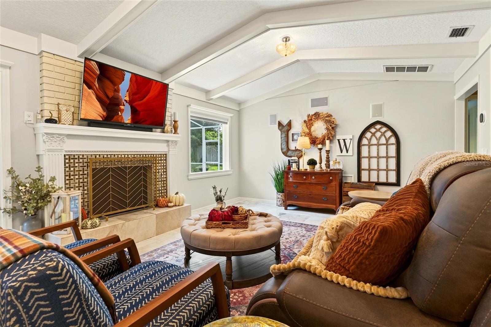 Furnished living room with a tiled fireplace, vaulted beamed ceiling, and large round ottoman.