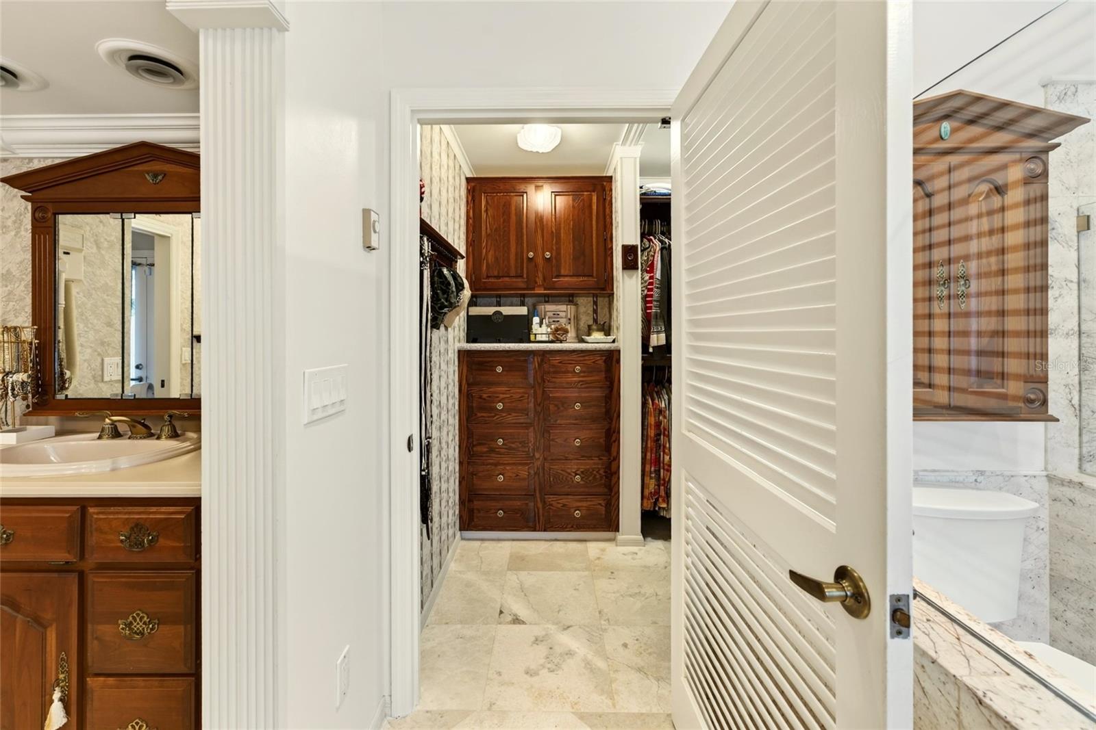 Bathroom vanity beside a walk-in closet with built-in wooden cabinets and marble-look tile.
