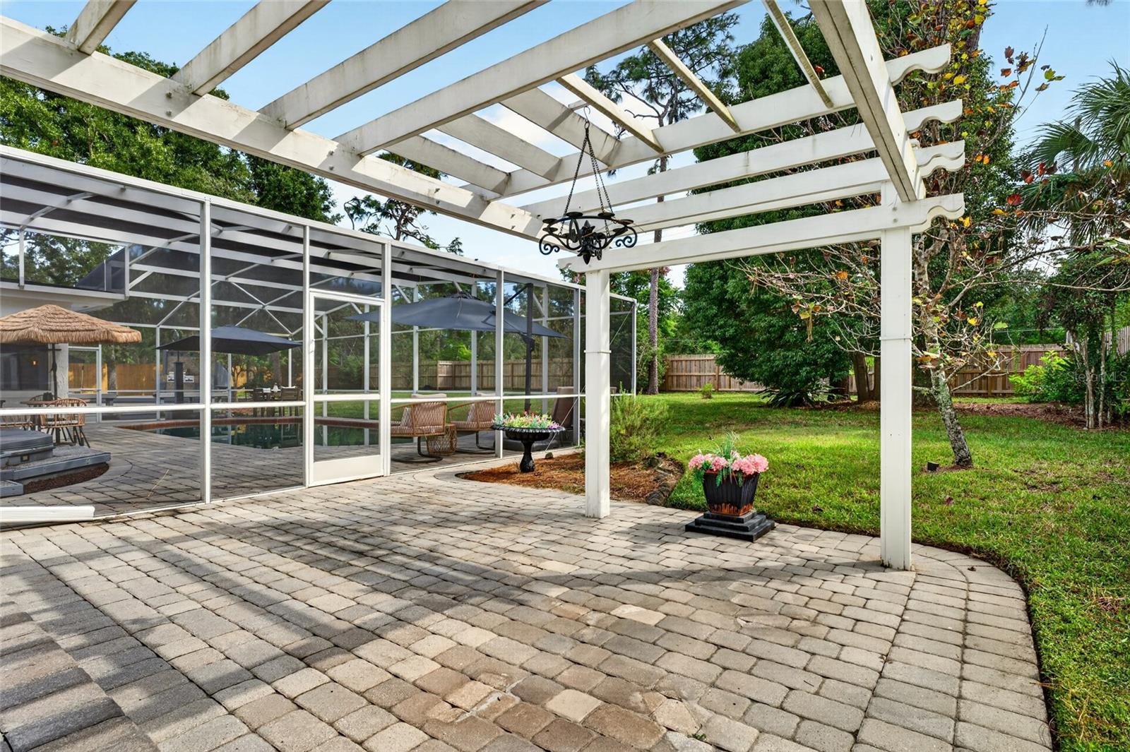 Paver patio beneath a white pergola beside a screened pool and landscaped backyard.