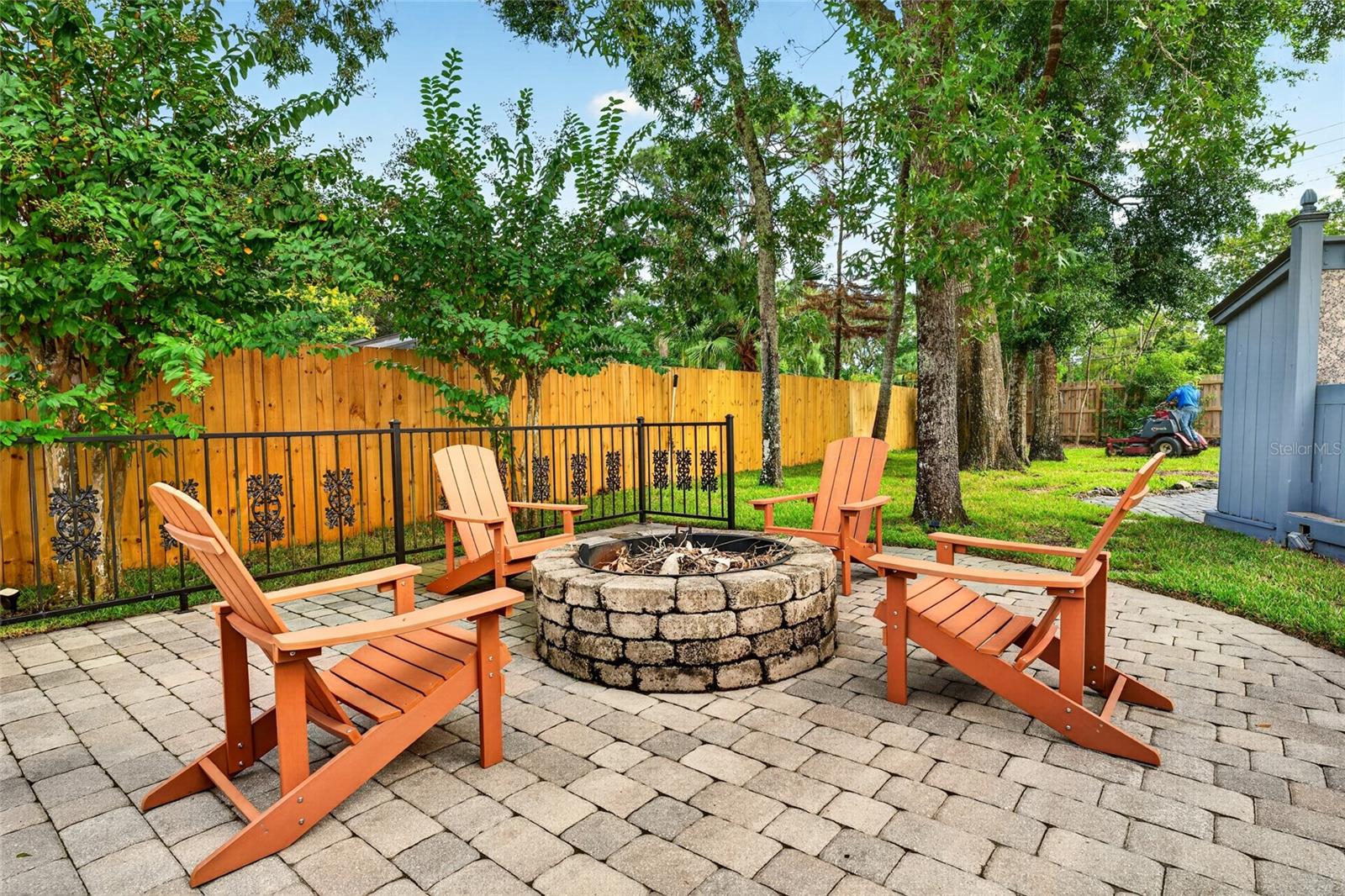 Backyard patio with a stone fire pit, orange Adirondack chairs, and mature shade trees.