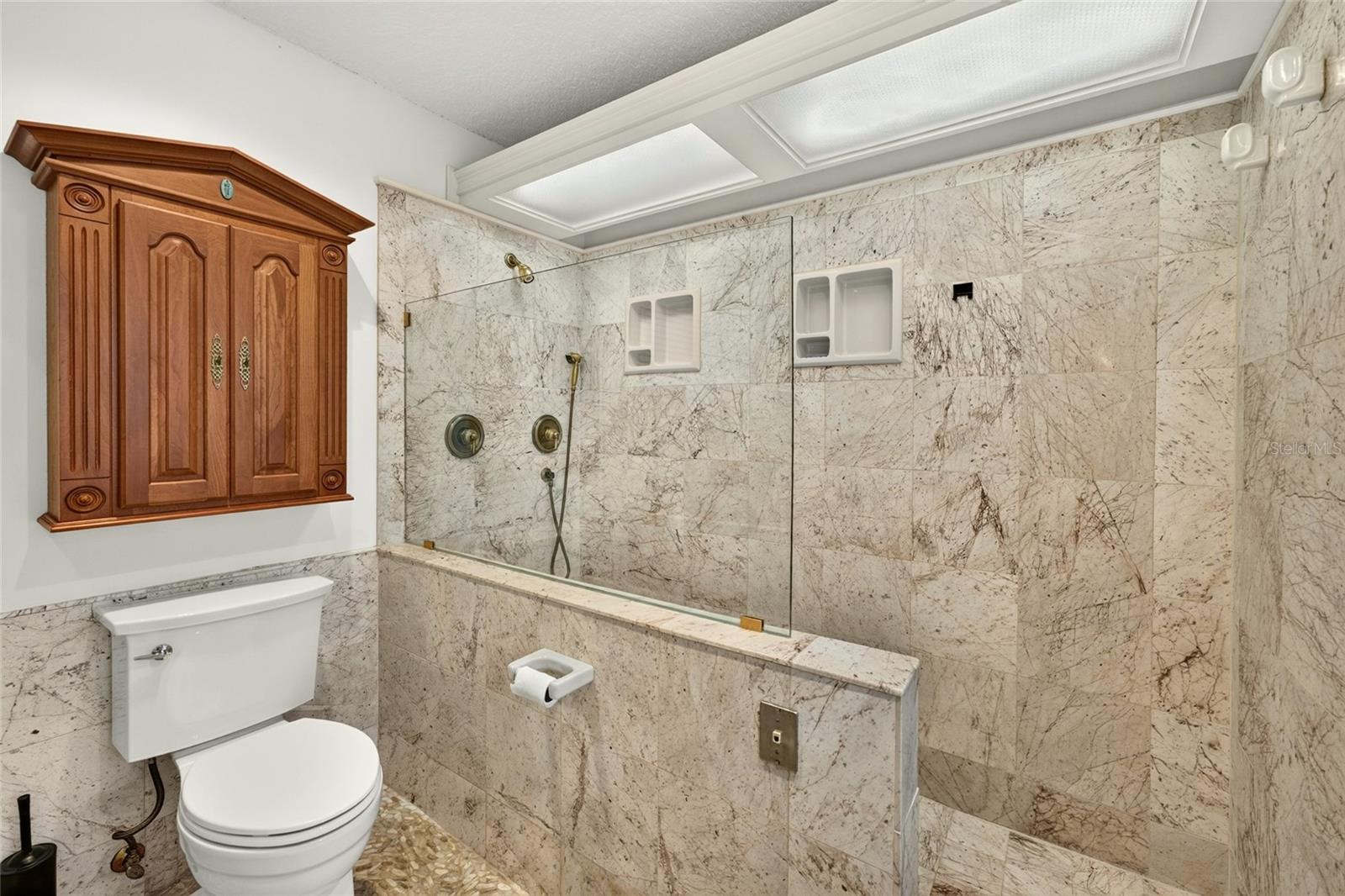 Marble-tiled bathroom with a glass-enclosed shower, brass fixtures, wooden wall cabinet, and white toilet.