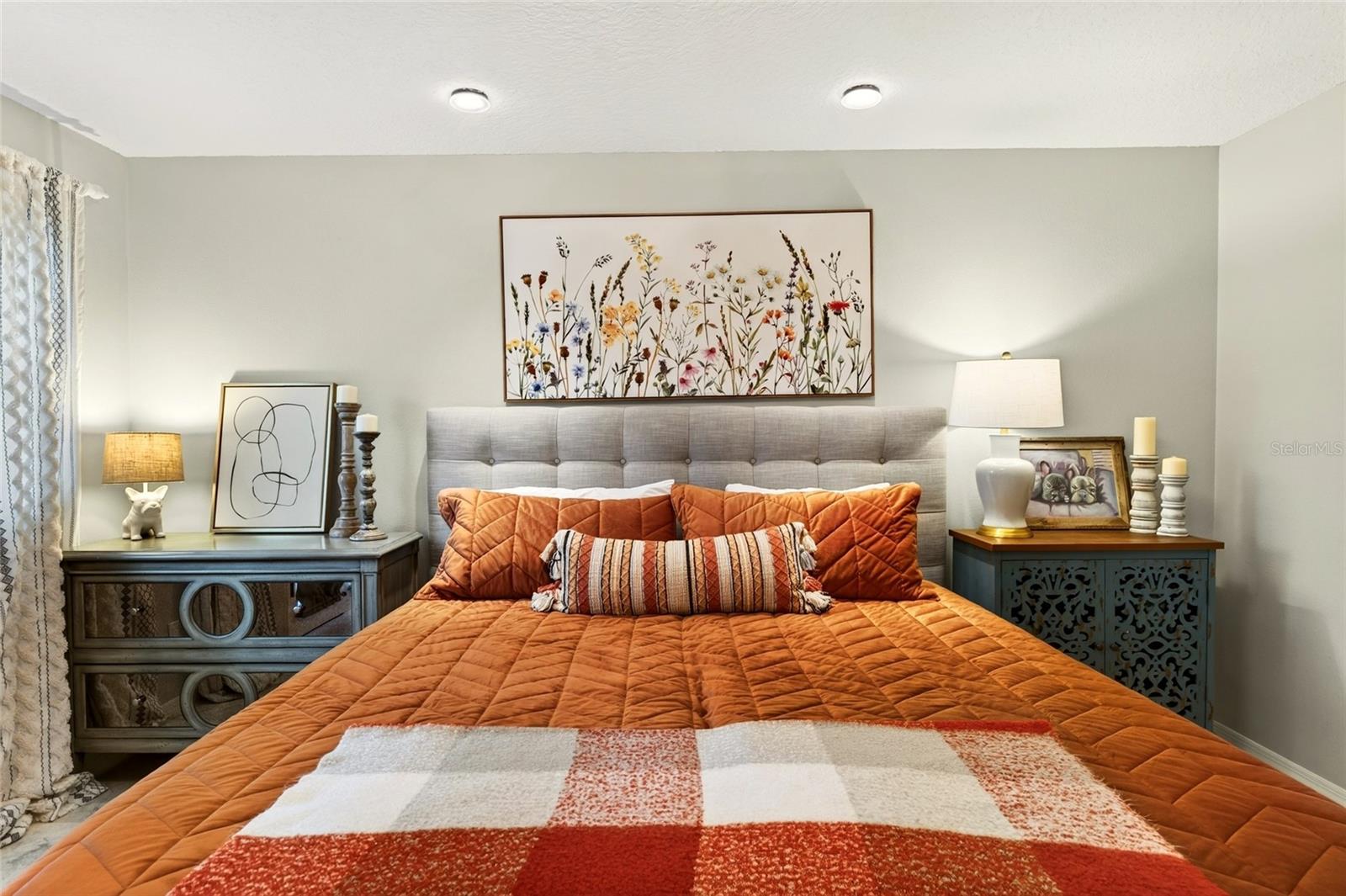 Bedroom with an orange quilted bed, gray upholstered headboard, and floral artwork.