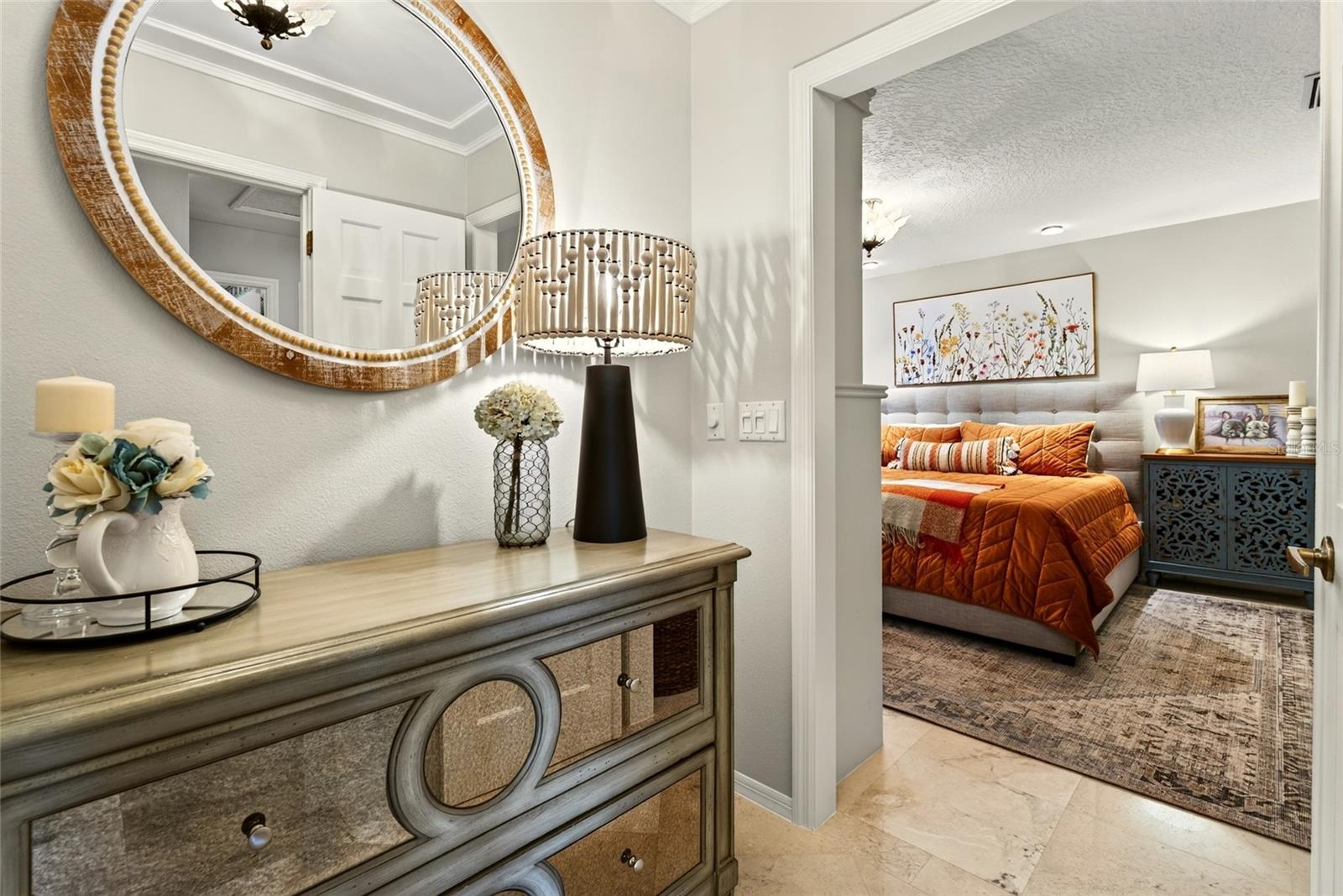 Furnished bedroom with an orange quilted bedspread, seen through a doorway beside a dresser and round mirror.