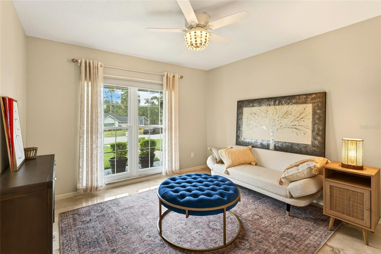 Furnished living room with a cream sofa, blue round ottoman, and large curtained window.