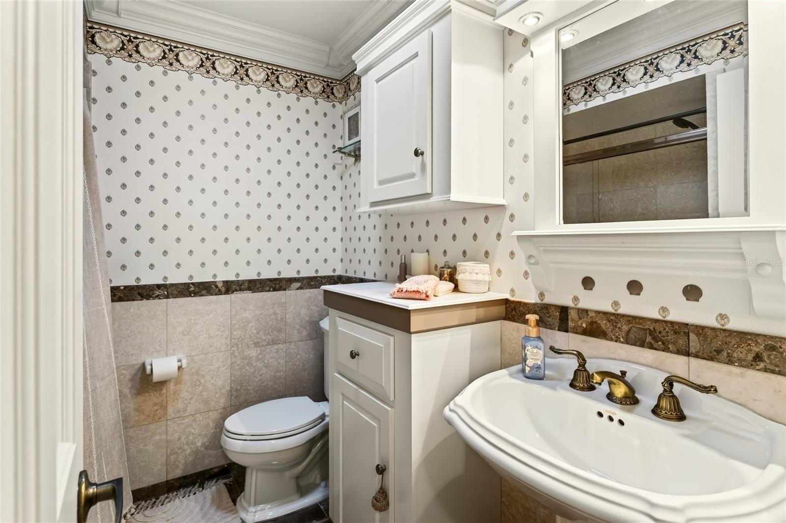 Bathroom with a curved white sink, toilet, large mirror, and patterned wallpaper.