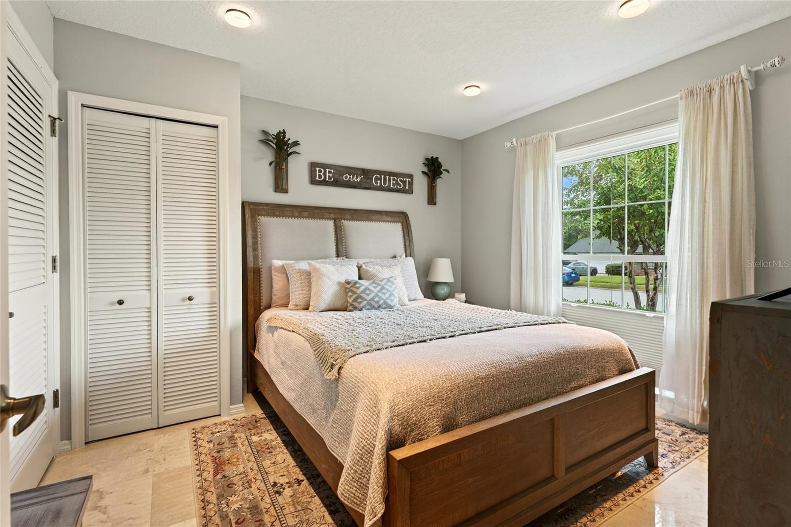 Guest bedroom with a wooden bed, neutral bedding, louvered closet doors, and a large curtained window.