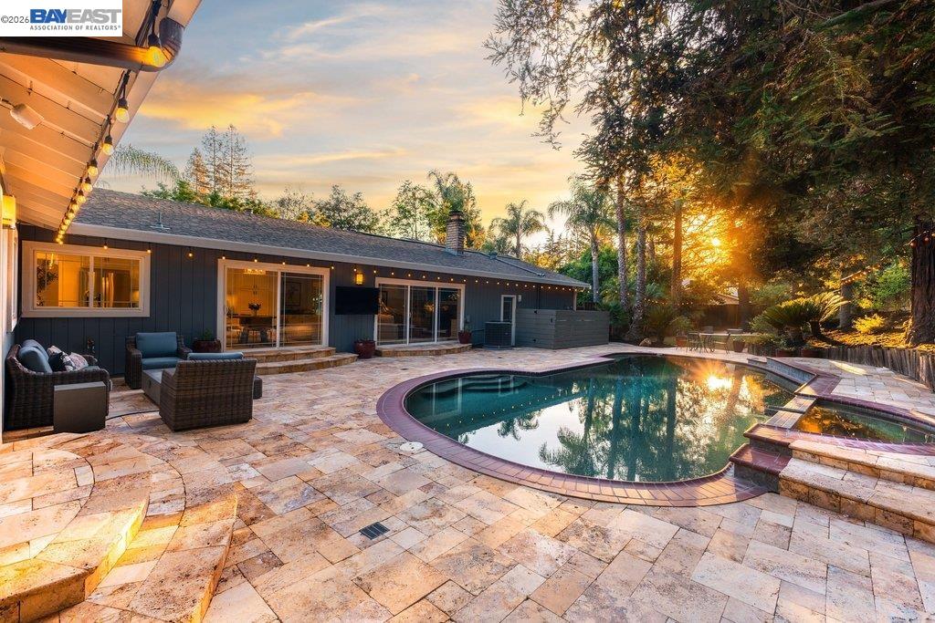 Backyard swimming pool beside a dark-sided home, with a stone patio and outdoor seating at sunset.