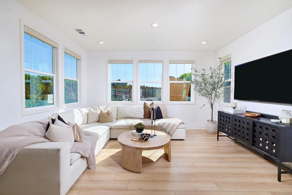 Living room with a white sectional, large windows, light wood floors, and a wall-mounted television.